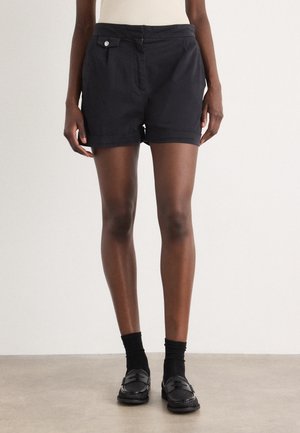 Personne portant un short noir taille haute avec détail de boutons, des chaussettes noires et des mocassins noirs, debout devant un fond clair uni.