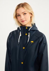 Ung kvinna med kort blont hår iförd en marinblå regnkappa med gula knappar och en paraplylogotyp, samt vita dragskosnören på huvan.