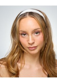 Ung kvinde med lysebrunt bølget hår iført et hvidt pandebånd og guldøreringe, der kigger direkte ind i kameraet med en neutral udtryk.