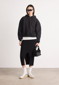 Haut à capuche noir surdimensionné, superposé sur une chemise blanche. Jupe en tricot côtelé noir avec une fente sur le côté. Portée avec des chaussettes blanches et des baskets argentées.