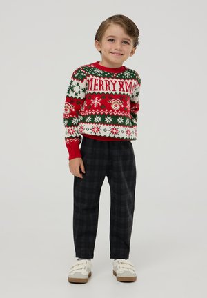 Maglione lavorato a maglia rosso, verde e bianco con la scritta "BUON NATALE", decorato con fiocchi di neve e una renna. Pantaloni a quadri scuri e scarpe bianche.