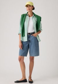 Άτομο με κίτρινο καπέλο, πράσινο ριγέ πουκάμισο πάνω από λευκή μπλούζα, τζιν σορτς μέχρι το γόνατο, μαύρη ζώνη, πράσινο πουλόβερ περασμένο στους ώμους και μαύρα μπαλαρίνια.