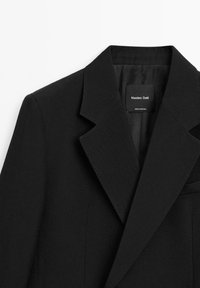 Černý sartorialní blazer s ostrým klopovým límcem, vykrojenými rameny a jednou kapsou na hrudi. Vyroben z texturované tkaniny.