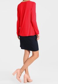 Woman wearing a red long-sleeve top, black knee-length skirt, and pink high-heeled shoes, standing with legs crossed, facing away.