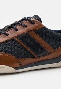 Men's sneaker featuring a navy mesh upper, brown leather accents, black logo patch, and contrasting stitching. Rubber sole with textured pattern.