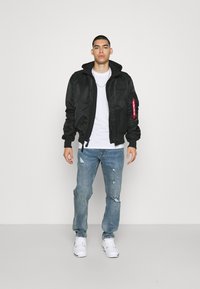 Schwarze Bomberjacke mit Kapuze, ausgestattet mit einem roten Reißverschluss und einer Vorderseite-Tasche, getragen über einem weißen T-Shirt und hellblauen, abgetragenen Jeans.