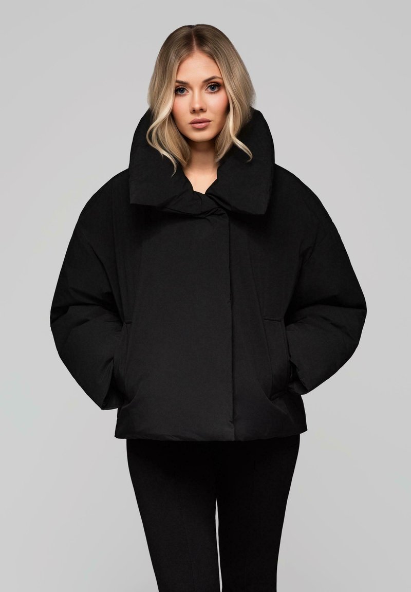 Blondýnka oblečená v černé oversized péřové bundě s širokým vysokým límcem a rukama v kapsách, stojící proti světle šedému pozadí.