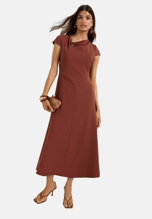 Nő barna midi ruhában, drapírozott nyakkivágással, fa borítéktáskát tart a kezében, barna magassarkú szandált és karkötőket visel.