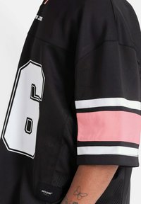 Camiseta deportiva negra con el número 6 en blanco. Presenta franjas rosas y blancas en las mangas. Hecha de una tela ligera con textura de malla. Etiqueta de marca visible.