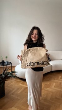 Brown crumpled paper bag with "zalarindo" printed in bold black letters, held by a person in a black top, standing in a bright room.