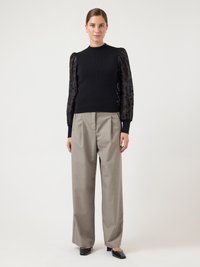 Woman standing wearing a black top with sheer floral sleeves, beige wide-leg trousers, and black square-toe heels on a white background.