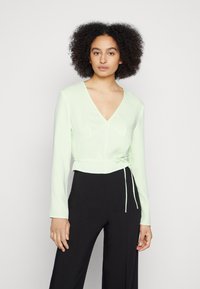 Blusa de manga longa verde-claro, com decote em V, cintura ajustada e fecho de cordão. Textura suave, design justo, combinada com calças pretas.