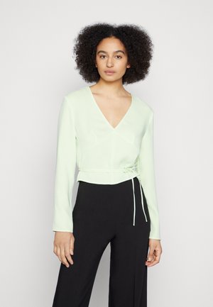 Blusa de manga longa verde-claro, com decote em V, cintura ajustada e fecho de cordão. Textura suave, design justo, combinada com calças pretas.