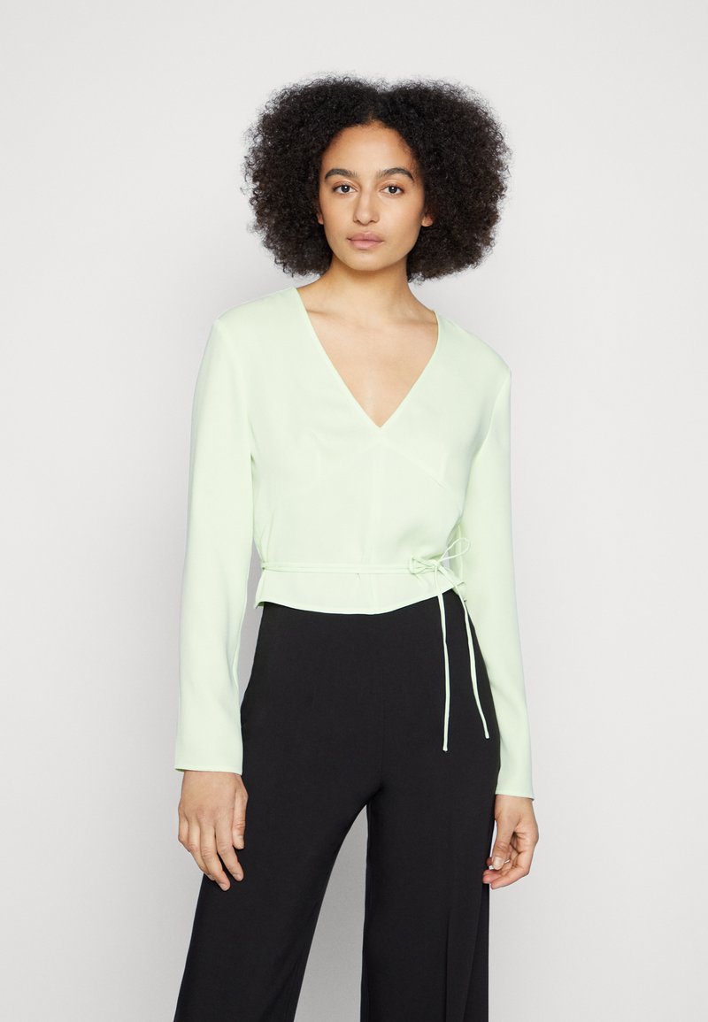 Blusa de manga longa verde-claro, com decote em V, cintura ajustada e fecho de cordão. Textura suave, design justo, combinada com calças pretas.