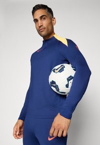 Mann i blå og gul sportsdrakt holder en hvit og blå fotball under armen, stående mot en enkel lys bakgrunn.