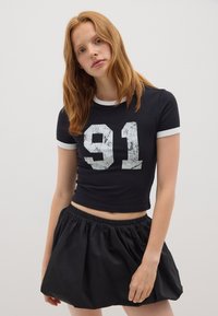 Top corto nero a maniche corte con dettagli bianchi, caratterizzato da una grafica grigia "91" consumata. Abbinato a una gonna nera leggera e gathering.