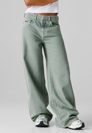 Inimene, kes kannab helerohelisi laia säärega teksapükse ja valgeid tossusid, seisab käed rüpes ühevärvilise tausta ees.