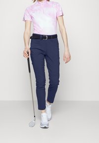 Jasnoróżowa koszulka z krótkim rękawem w stylu tie-dye z zamkiem, w zestawie z ciemnoniebieskimi, dopasowanymi spodniami golfowymi, białymi butami sportowymi i kijem golfowym.