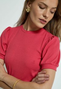 Růžový žebrovaný top s krátkým rukávem, kulatým výstřihem a nadýchanými rukávy, doplněný zlatými náušnicemi ve tvaru květiny, nošený modelem.