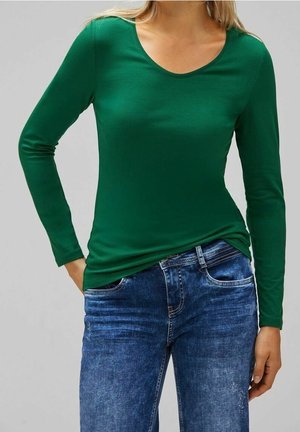 Femme portant un t-shirt vert ajusté à manches longues et un jean bleu en denim, posant avec une main dans la poche devant un fond gris uni.