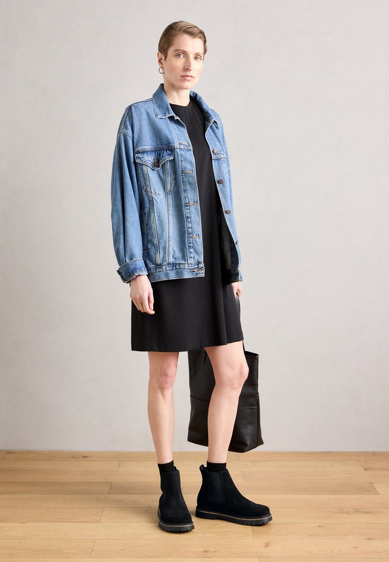 Veste en denim, bleu clair, coupe oversize, fermeture à boutons ; robe noire, longueur genoux ; bottines noires en daim, à la cheville ; sac fourre-tout noir.