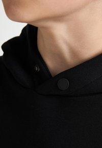 Col montant d'un hoodie noir avec un tissu texturé, une capuche proéminente, et une fermeture à pression ornée d'un logo rond.