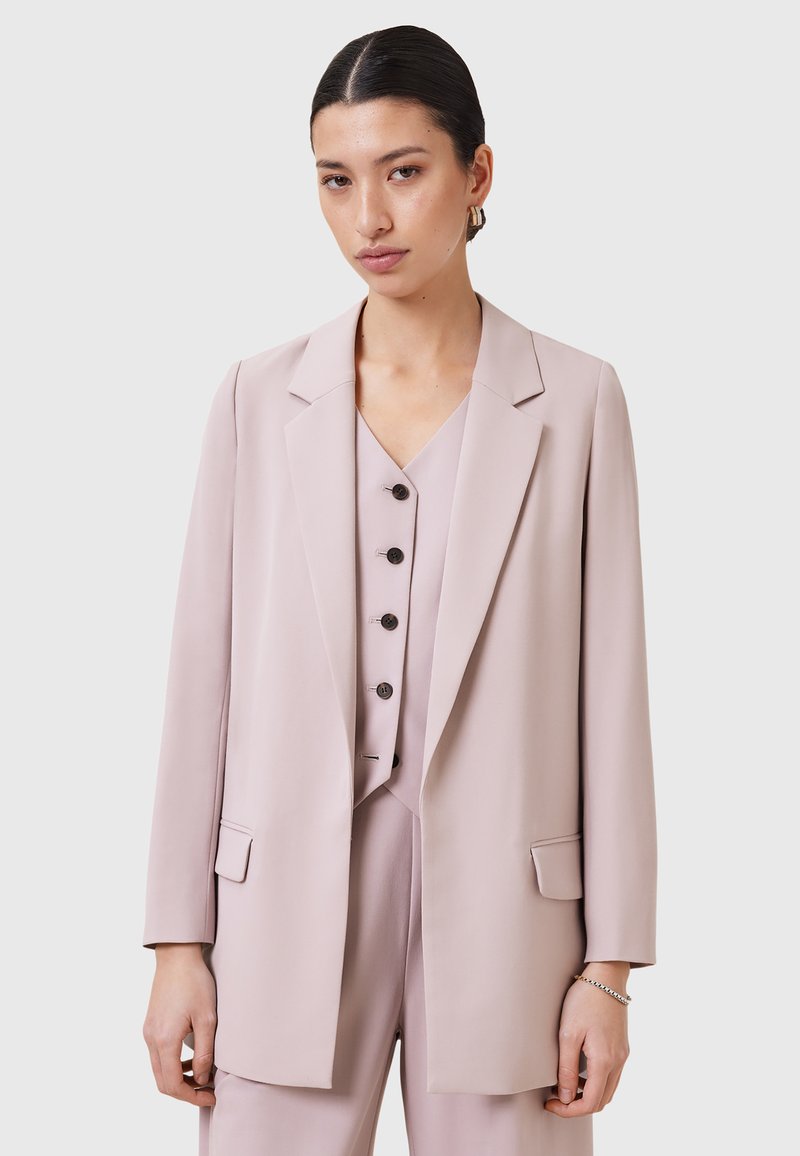 Blazer su misura rosa chiaro con maniche lunghe, revers a punta e tasche. Indossato sopra una camicetta coordinata con cinque bottoni e scollatura a V.