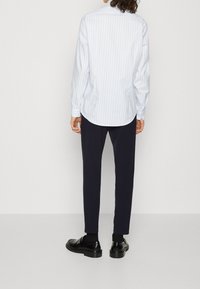 Camisa de rayas azul claro y blanca con mangas largas, dobladillo curvado, puños con botones, combinada con pantalones oscuros entallados y zapatos negros.