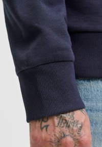 Dark blue sweater cuff displaying ribbed texture and seamless design, partially covering a tattooed hand. Material appears soft and stretchy.