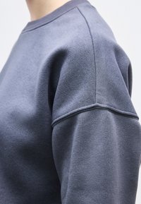 Κοντινό πλάνο ενός ατόμου που φοράει γκρι πουκάμισο με μακριά μανίκια, εστιάζοντας στις λεπτομέρειες των ραφών στον ώμο και στο άνω μέρος του μπράτσου.