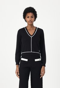 Maglione nero a V con bordi bianchi, maniche lunghe e design aderente. Abbinato a pantaloni neri a trama con accenti bianchi sulle tasche.