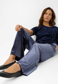 Top en sequins bleu marine associé à un pantalon large en velours bleu clair et des chaussures plates noires. Position assise avec les jambes étendues vers l'avant.