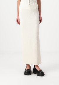 Fustă midi tricotată, albă, cu un model texturat, având un tiv fitat și un tiv ondulat. Asortată cu pantofi slip-on negri, voluminoși.