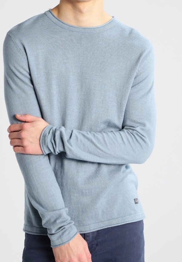 Homme portant une chemise bleu clair à manches longues avec un petit logo près de l'ourlet, croisant un bras sur l'autre devant un fond uni.