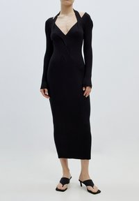 Schwarzes geripptes Strickkleid mit langen Ärmeln, tiefem V-Ausschnitt und taillierter Silhouette. Kombiniert mit schwarzen Sandalen mit offenen Zehen vor einem hellen Hintergrund.