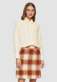 Crème blouse met een strikkraag gecombineerd met een knielange geruite rok met rode, oranje en witte vierkanten. Zachte, gestructureerde stof.