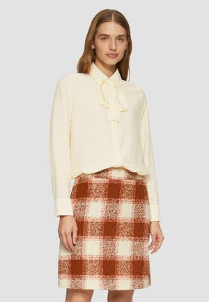 Crème blouse met een strikkraag gecombineerd met een knielange geruite rok met rode, oranje en witte vierkanten. Zachte, gestructureerde stof.