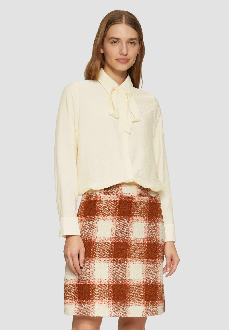 Crème blouse met een strikkraag gecombineerd met een knielange geruite rok met rode, oranje en witte vierkanten. Zachte, gestructureerde stof.