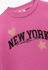 Roze sweatshirt met geribde kraag met de tekst "NEW YORK Manhattan 1983". Versierd met een geruit hart en ster motief.