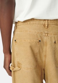 Pantalones de mezclilla beige con cintura alta, dos bolsillos traseros y un detalle de lazo en el lateral. La tela tiene una textura suave con costuras visibles.