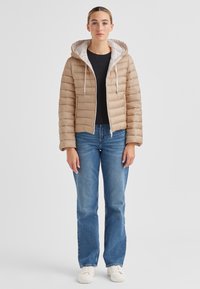 Steppjacke in Hellbraun mit weißem Futter und Kapuze, ausgestattet mit einem Reißverschluss und kastiger Form. Kombiniert mit blauen, gerade geschnittenen Jeans und weißen Schuhen.