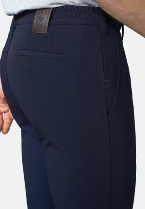 Nahaufnahme einer Person, die marineblaue Hosen mit elastischem Bund und einem Lederpatch mit der Aufschrift "M5" über der Gesäßtasche trägt.