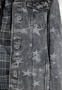Grijze denimjas met lichte sterpatronen over een zwart-wit geblokte hoodie met zichtbare rits en koordjes.