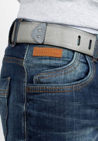 Jeans en jean bleu avec des coutures orange et une étiquette en cuir portant la mention "CIPO & BAXX denim". Une ceinture en cuir gris avec des accents découpés.