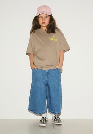 Dziecko z długimi brązowymi włosami, ubrane w różową czapkę, beżowy, luźny t-shirt, szerokie niebieskie dżinsy, białe skarpetki oraz czarno-białe slipony w kratkę.