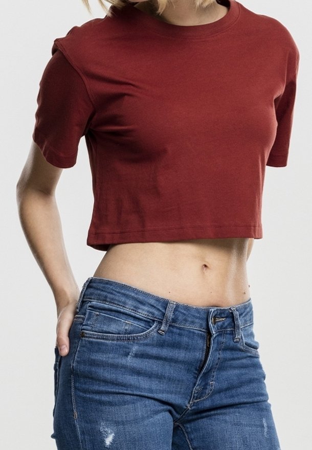 Kvinde iført en tætsiddende rød cropped t-shirt og blå denim jeans, stående med den ene hånd i lommen mod en ensfarvet baggrund.