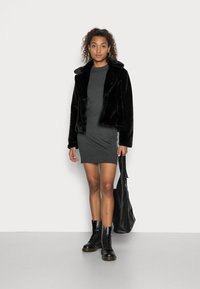 Veste en fausse fourrure noire, robe grise, bottes de combat noires, tenant un sac fourre-tout noir ; texture lisse et coupe ajustée.