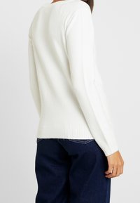 Vit stickad tröja med långa ärmar, ribbade mudd och nederkant; mjuk textur och minimalistisk design. Buren med mörkblå jeans.