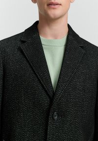 Zwarte geweven blazer met een visgraatpatroon, met een enkele knoopsluiting en notched revers, over een lichtgroene trui met een ronde hals.