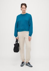 Teal sweatshirt met verlaagde schouders, gecombineerd met lichtbeige joggingsbroek. Zwarte scrunch tas in één hand, en grijze sneakers aan.